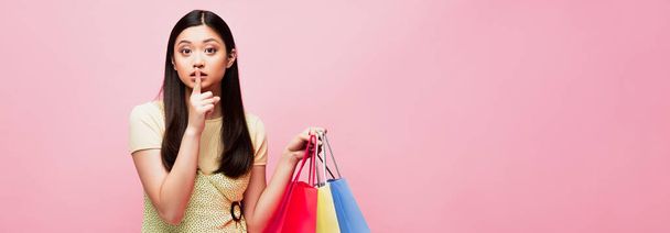 vízszintes koncepció ázsiai lány nézi a kamera, miközben gazdaság bevásárlószatyrok és bemutató csend jel elszigetelt rózsaszín  - Fotó, kép