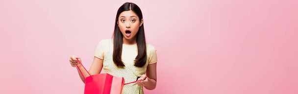panoramic crop of shocked young asian woman holding shopping bag on pink  - Photo, Image