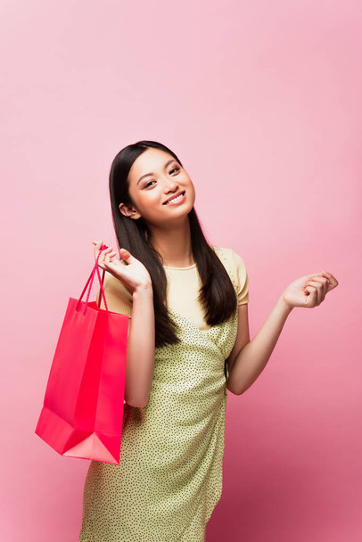 smiling young asian woman holding shopping bag on pink  - Fotó, kép