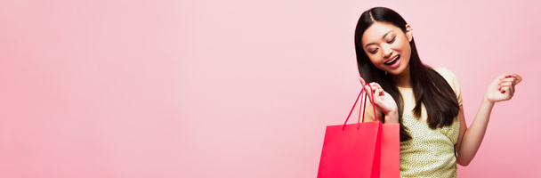 panoramic crop of smiling young asian woman looking at shopping bag on pink  - Фото, изображение