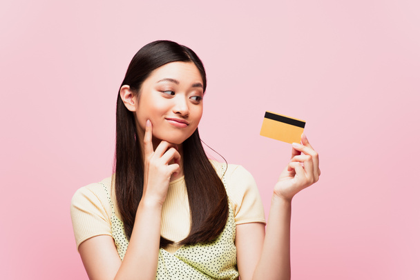 pensive asian woman looking at credit card isolated on pink  - Foto, Imagem
