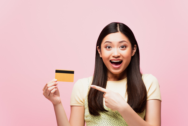 excited asian woman pointing with finger at credit card isolated on pink  - Fotó, kép