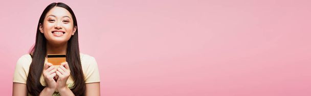 horyzontalny obraz wesołej i młodej azjatki posiadającej kartę kredytową odizolowaną na różowo  - Zdjęcie, obraz