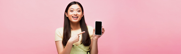 cultivo panorámico de mujer asiática positiva apuntando con el dedo al teléfono inteligente con pantalla en blanco aislado en rosa  - Foto, Imagen