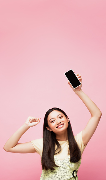joyeuse fille asiatique avec poing serré tenant smartphone avec écran vide isolé sur rose  - Photo, image