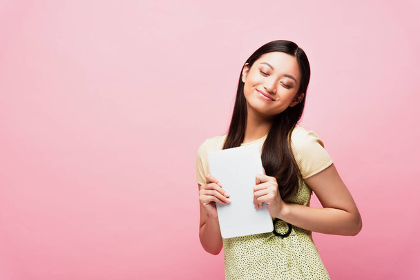 cheerful asian girl with closed eyes holding digital tablet isolated on pink  - Fotó, kép