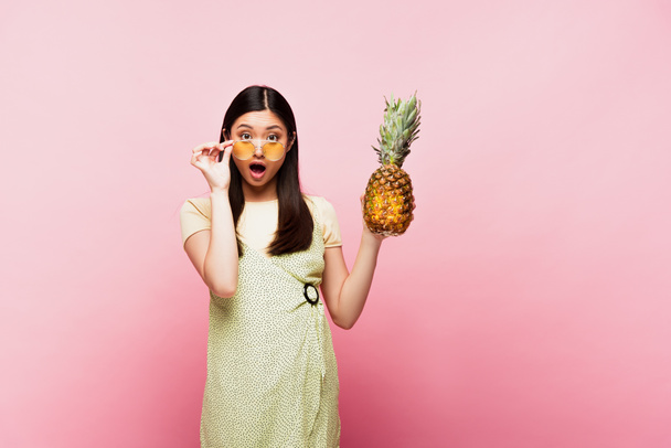 choqué asiatique fille dans lunettes de soleil tenue mûr ananas sur rose  - Photo, image