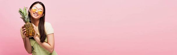 cosecha horizontal de feliz asiático chica en gafas de sol celebración maduro piña aislado en rosa  - Foto, Imagen