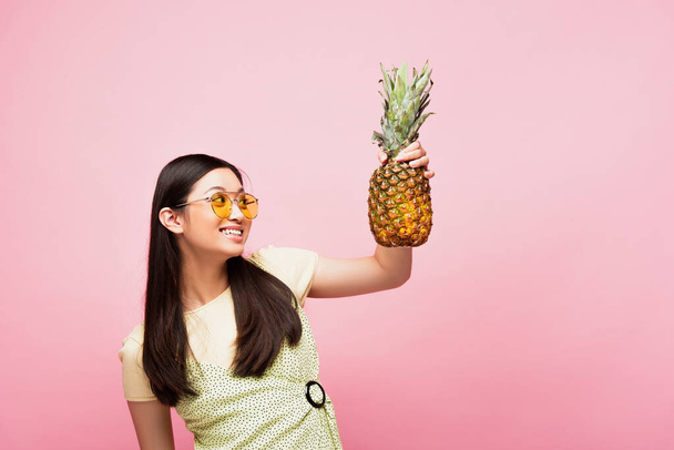 smiling asian woman in sunglasses looking at fresh pineapple isolated on pink  - Фото, зображення