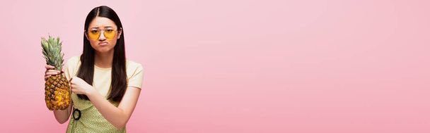 concepto horizontal de mujer asiática triste en gafas de sol sosteniendo piña fresca aislada en rosa  - Foto, Imagen