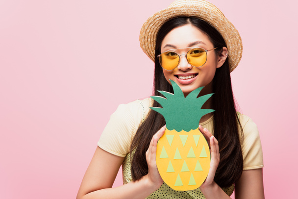 веселая азиатская женщина в солнечных очках и соломенной шляпе с бумажным ананасом, изолированным на розовом  - Фото, изображение