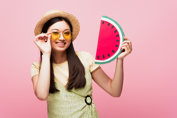 vidám ázsiai lány szalma kalap megható napszemüveg és gazdaság papír görögdinnye elszigetelt rózsaszín  - Fotó, kép