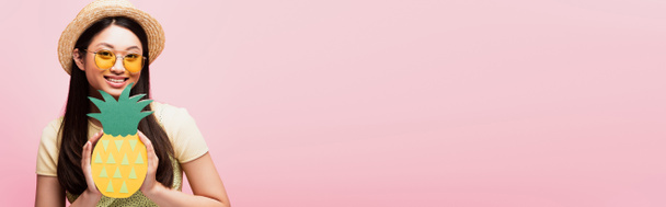 cultivo panorámico de alegre chica asiática en gafas de sol y sombrero de paja sosteniendo papel piña aislado en rosa  - Foto, Imagen