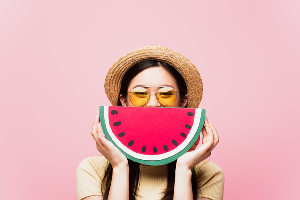 asian girl in sunglasses and straw hat covering face with paper watermelon isolated on pink  - Photo, Image