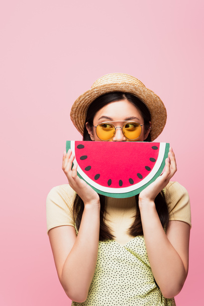 Asiatin mit Sonnenbrille und Strohhut verdeckt Gesicht mit Papierwassermelone und schaut isoliert auf rosa  - Foto, Bild
