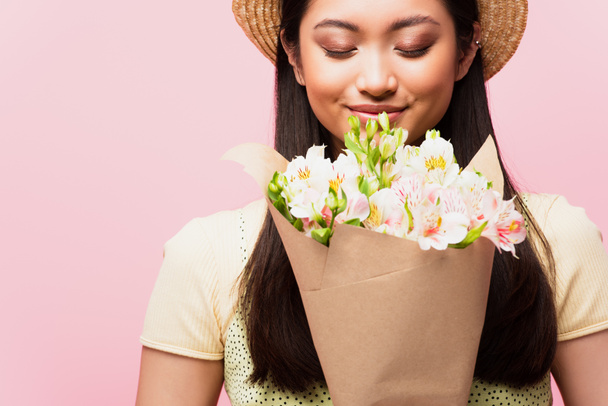positive asian girl in straw hat smelling flowers isolated on pink  - Photo, Image