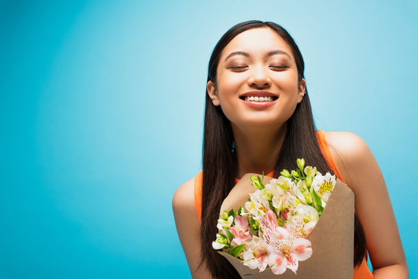 happy asian girl with closed eyes smiling and holding flowers on blue - Fotó, kép
