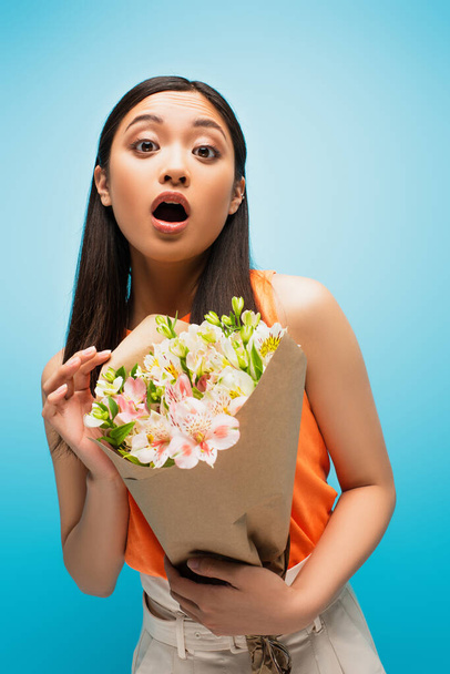 surprised asian girl looking at camera and holding flowers on blue - Foto, afbeelding