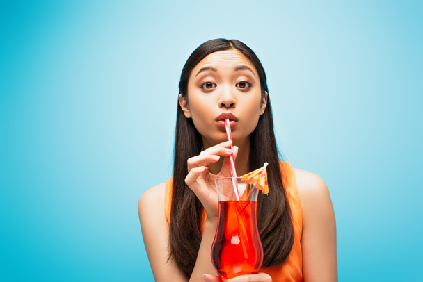 atractivo asiático chica celebración de vidrio y beber cóctel a través de paja en azul - Foto, Imagen