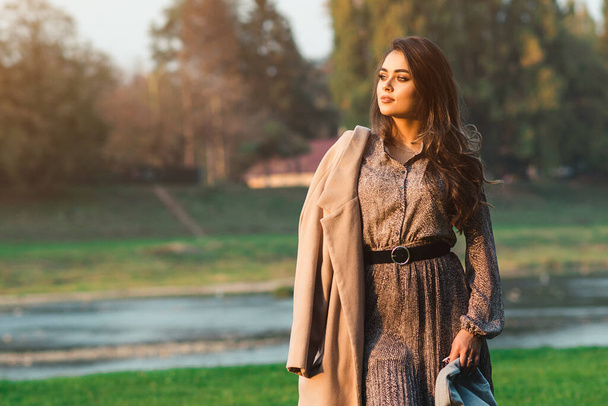 Fashionable woman walking in city park. Beauty and fashion. Stylish girl wearing trendy dress and coat. Woman with perfect makeup and hairstyle. Autumn fashion, lifestyle. - Photo, Image
