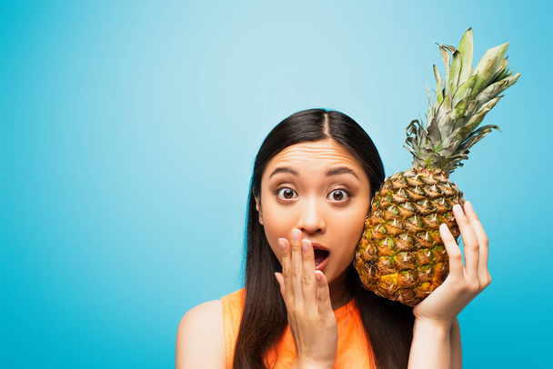 шокована азіатська дівчина покриває рот і тримає стиглий ананас на синьому
  - Фото, зображення