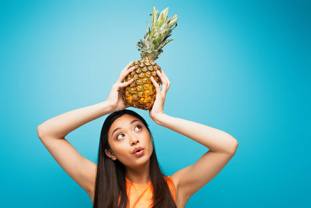 beautiful asian girl holding ripe pineapple above head on blue  - Photo, Image
