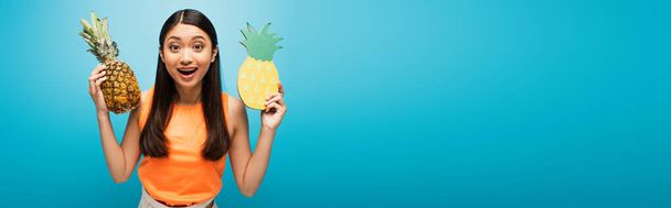 panoramic concept of excited asian girl holding ripe and carton pineapples on blue  - 写真・画像