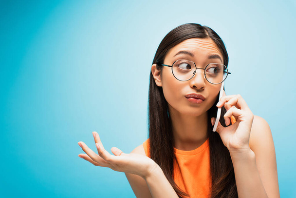 attractive asian girl in glasses talking on smartphone and gesturing on blue - Fotografie, Obrázek