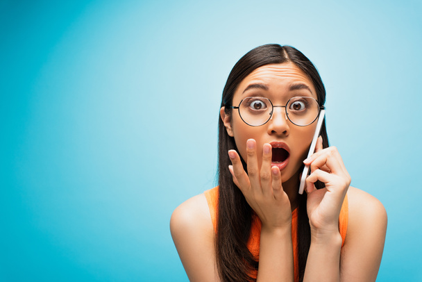 shocked asian girl in glasses covering mouth while talking on smartphone on blue - Zdjęcie, obraz