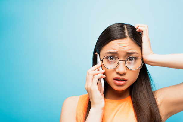 confus asiatique fille dans lunettes parler sur smartphone et toucher la tête sur bleu - Photo, image