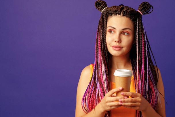 Young caucasian woman with colorful long cornrows holding a cup of coffee against purple background - Φωτογραφία, εικόνα