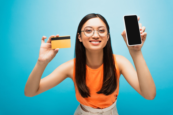 happy asian girl holding smartphone with blank screen and credit card on blue  - Foto, afbeelding