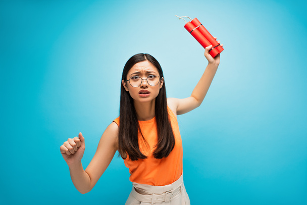 emotional asian woman in glasses holding dynamite sticks above head on blue - Photo, Image