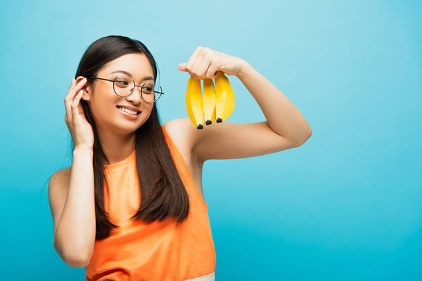 счастливая азиатская девушка в очках, глядя на спелые бананы, изолированные на голубом  - Фото, изображение