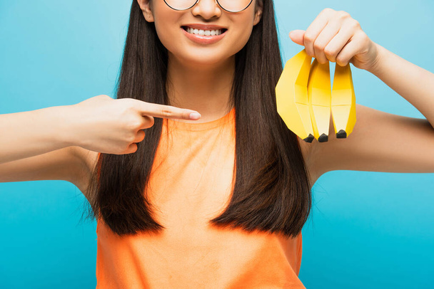 przycięty widok wesołej dziewczyny w okularach wskazujących palcem na dojrzałe banany odizolowane na niebiesko  - Zdjęcie, obraz