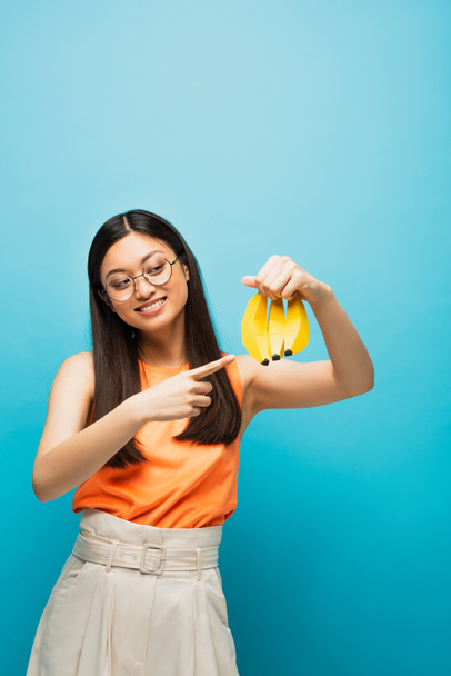 cheerful asian girl in glasses pointing with finger at ripe bananas isolated on blue  - 写真・画像