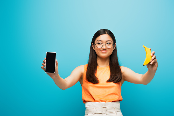 mujer asiática positiva en gafas sosteniendo teléfono inteligente con pantalla en blanco y plátano en azul  - Foto, imagen