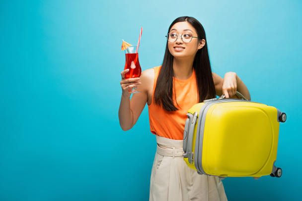 cheerful asian girl in glasses holding cocktail and baggage while looking away on blue  - Photo, Image