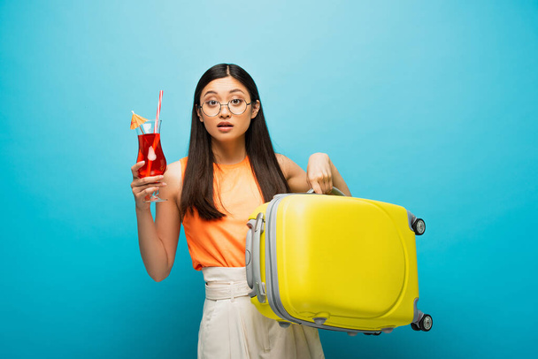 young asian woman in glasses holding baggage and cocktail on blue  - Фото, изображение