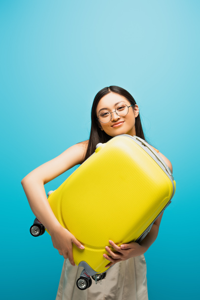 cheerful asian woman in glasses holding yellow luggage on blue  - Photo, Image