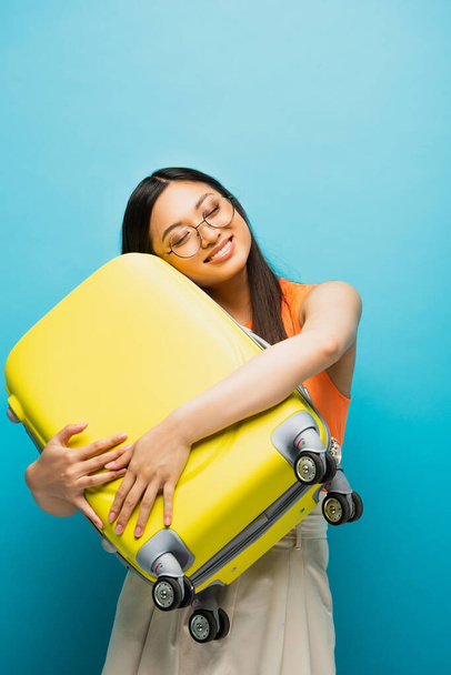 cheerful asian woman in glasses hugging yellow luggage on blue  - Fotó, kép