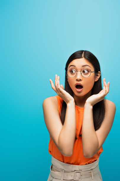 shocked asian woman in glasses gesturing and looking away isolated on blue - Φωτογραφία, εικόνα