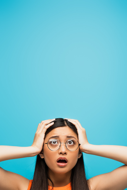 surprised asian woman in glasses touching head isolated on blue - Foto, afbeelding