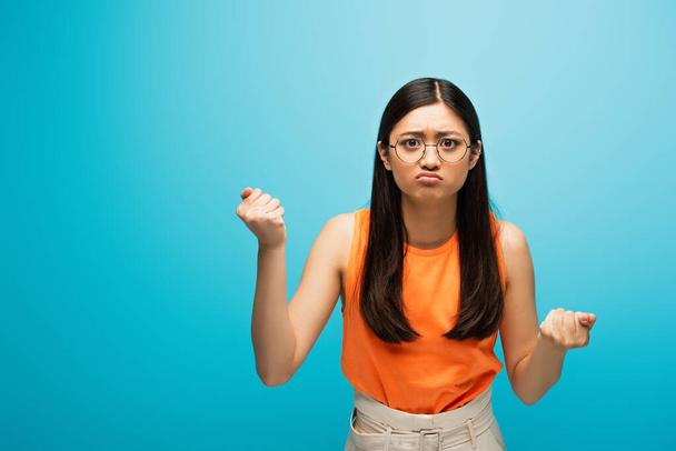 frustrated asian girl in glasses showing clenched fists on blue - 写真・画像