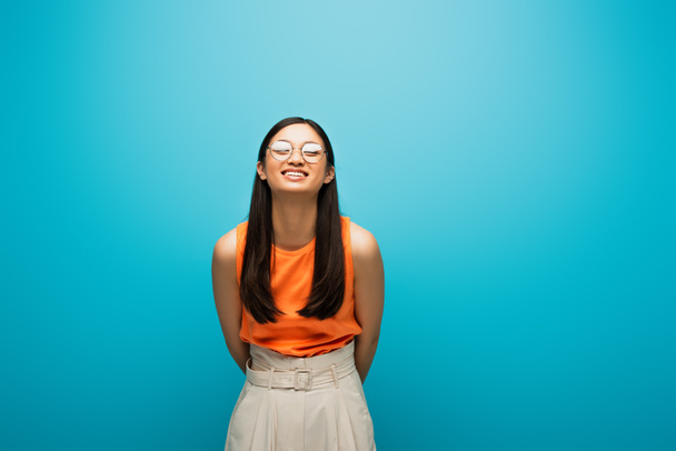 cheerful asian girl in glasses smiling and standing on blue - Zdjęcie, obraz