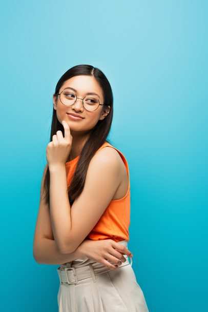 cheerful asian girl in glasses smiling and looking away isolated on blue - Valokuva, kuva