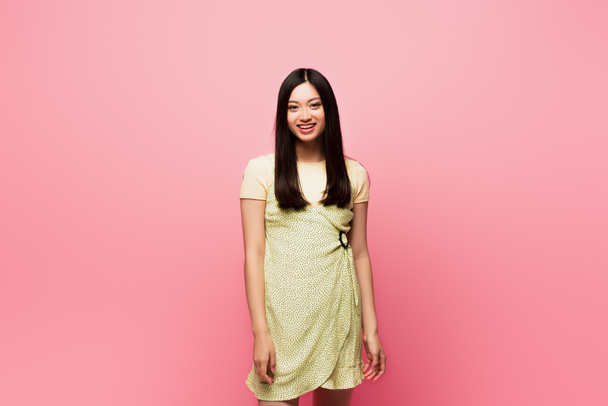 positive junge asiatische Frau blickt in die Kamera und lächelt auf rosa  - Foto, Bild
