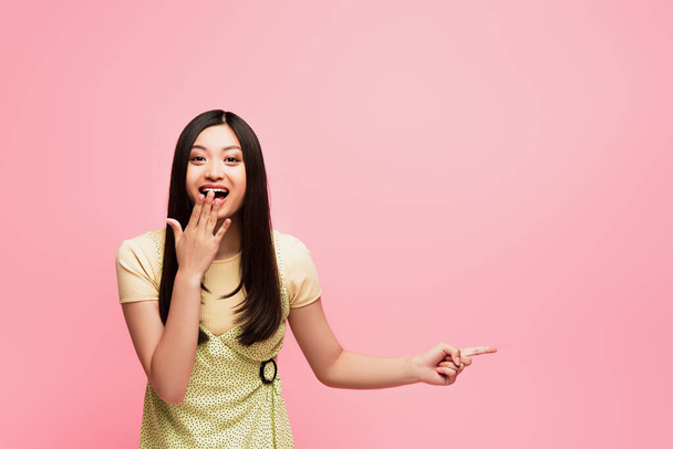 excited asian girl pointing with finger and covering mouth isolated on pink  - Photo, Image