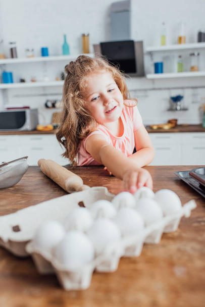 selective focus of blonde girl taking chicken egg near rolling pin on kitchen table - Фото, зображення
