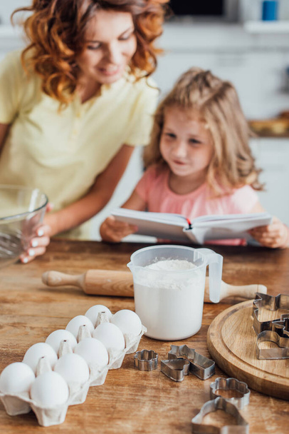 επιλεκτική εστίαση των αυγών κοτόπουλου, αλεύρι στη μέτρηση κανάτα και κόφτες μπισκότων κοντά στη μητέρα και το παιδί ανάγνωση βιβλίο μαγειρικής - Φωτογραφία, εικόνα
